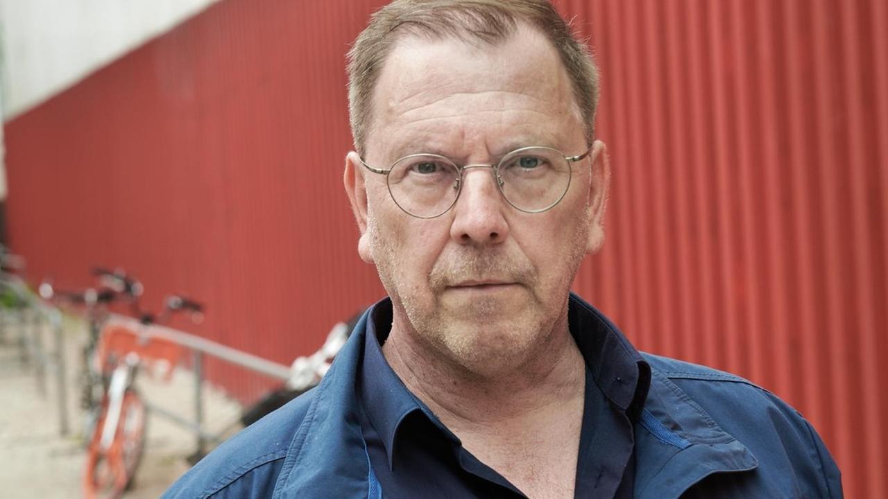 Ein Mann in blauem Hemd und blauer Jacke, mit Brille, schaut in die Kamera. Im Hintergrund ist ein Gebäude aus rotem und weißem Wellblech zu sehen. Ein Mann in blauem Hemd und blauer Jacke, mit Brille, schaut in die Kamera. Im Hintergrund ist ein Gebäude aus rotem und weißem Wellblech zu sehen.