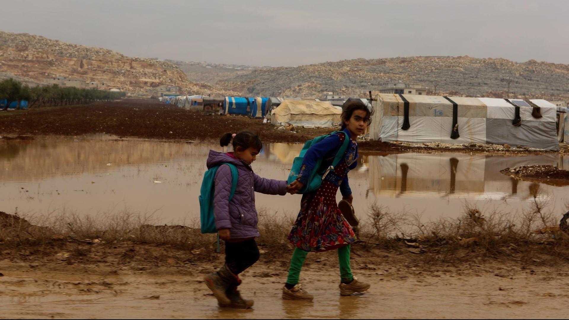Zwei Kinder laufen durch Matsch. Im Hintergrund sind weiße Flüchtlingszelte zu erkennen.