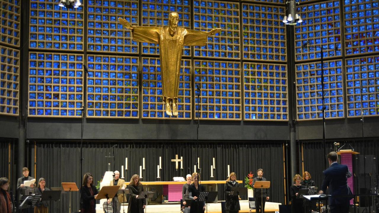 Ein Kammerchor steht im großen Abstand voneinander in dem modernen Kirchenraum vor einer großen, goldenen Jesusfigur in Kreuzform, die im Altarraum hängt.