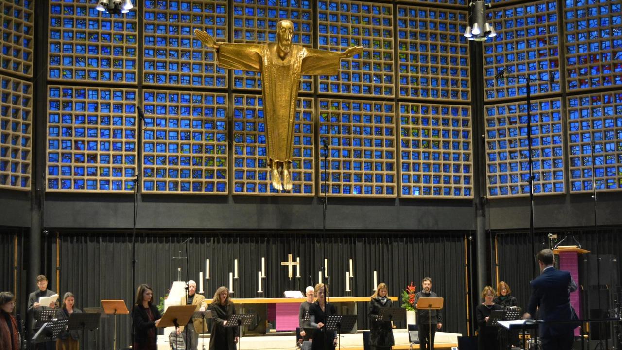 Ein Kammerchor steht im großen Abstand voneinander in dem modernen Kirchenraum vor einer großen, goldenen Jesusfigur in Kreuzform, die im Altarraum hängt. Ein Kammerchor steht im großen Abstand voneinander in dem modernen Kirchenraum vor einer großen, goldenen Jesusfigur in Kreuzform, die im Altarraum hängt.