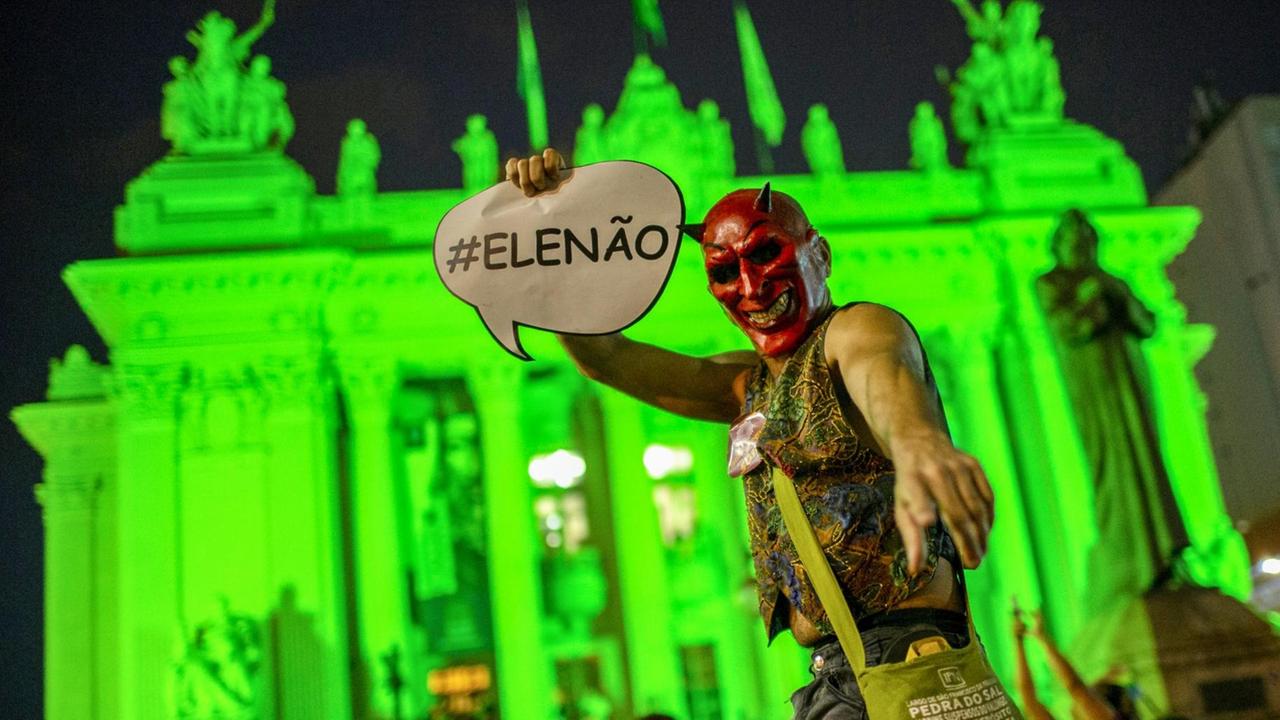 Ein Demonstrant mit Teufelsmaske hält ein Plakat mit "#EleNao" in die Kamera vor einem neongrün illuminierten historischen Gebäude.