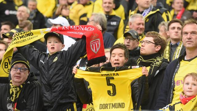 Dortmund-Fans auf der Tribüne. Ein Junge hält einen Fan-Schal mit Borussen- und Monaco-Logo.