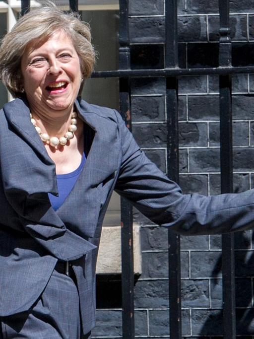 Theresa May geht lachend am Zaun eines Gebäudes vorbei
