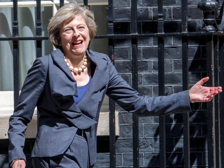 Theresa May geht lachend am Zaun eines Gebäudes vorbei