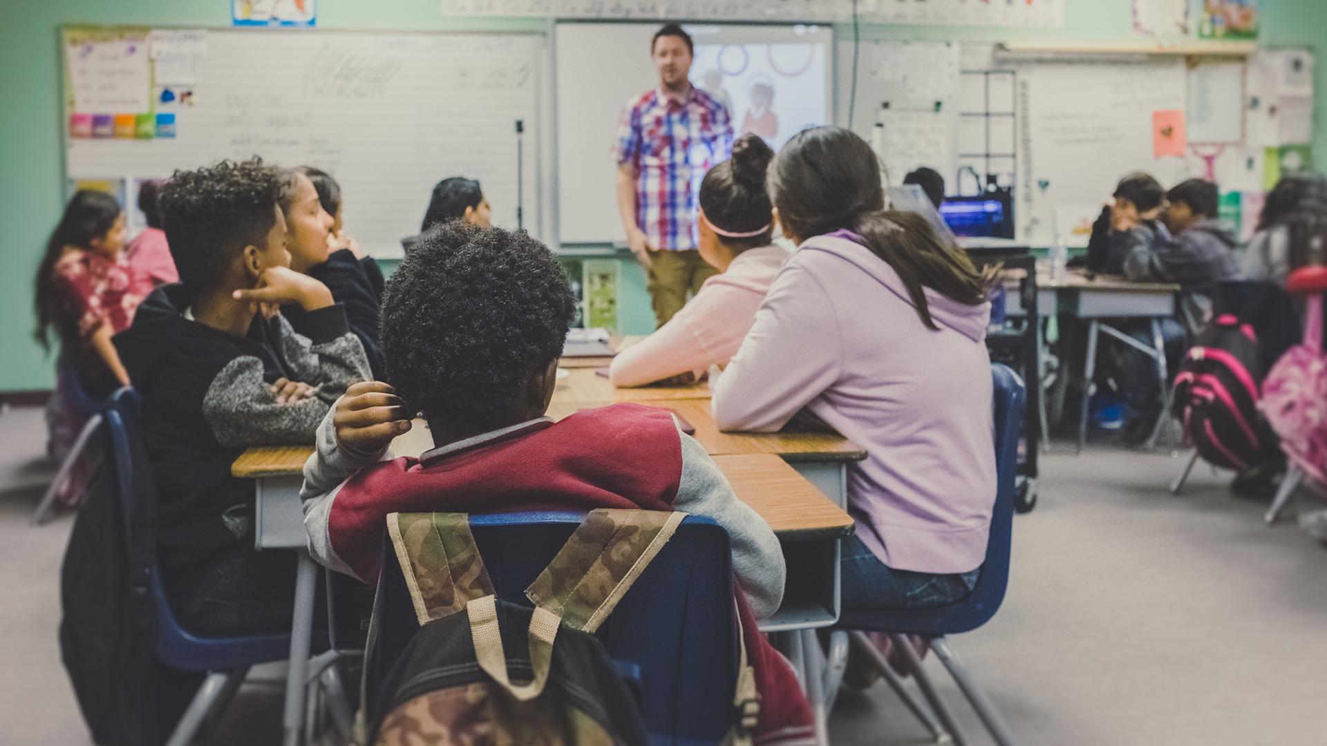 Ein Schülergruppe sitzt während des Unterrichts an einem Tisch - vorn steht der Lehrer und erklärt etwas. Ein Schülergruppe sitzt während des Unterrichts an einem Tisch - vorn steht der Lehrer und erklärt etwas.