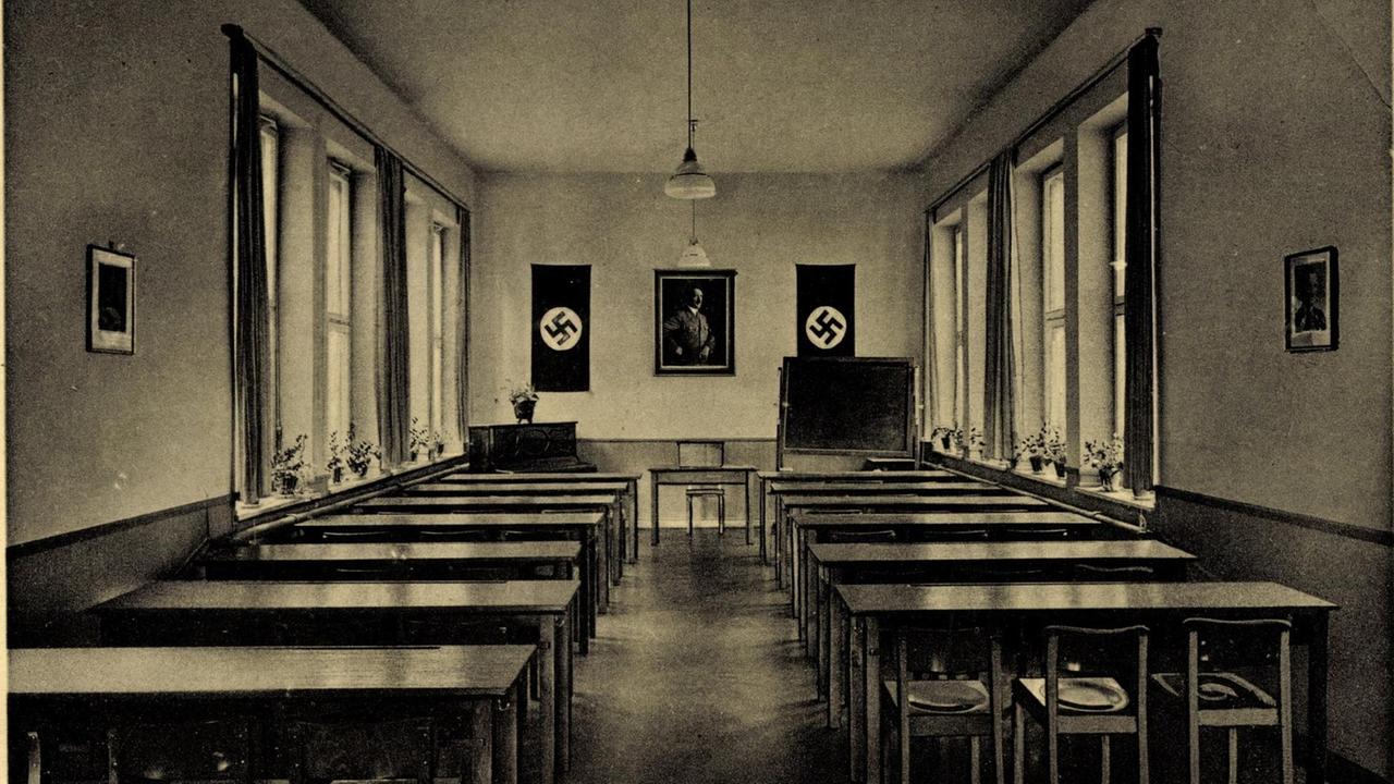 Eine Schwarzweißpostkarte zeigt in Zentralperspektive einen langgestreckten Sitzungssaal mit Reihen von Pulten jeweils an der rechten und der linken Seite des Raumes. Am Kopfende sind zwei Hakenkreuze und ein Führerbild, das Adolf Hitler abbildet, zu sehen.