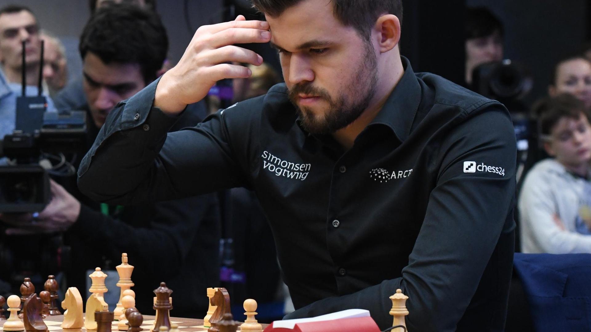 Schachweltmeister Magnus Carlsen mit kurzrasiertem Bart und schwarzem Hemd sitz vor einem Schachbrett und hält sich die Hand an den Kopf. Schachweltmeister Magnus Carlsen mit kurzrasiertem Bart und schwarzem Hemd sitz vor einem Schachbrett und hält sich die Hand an den Kopf.