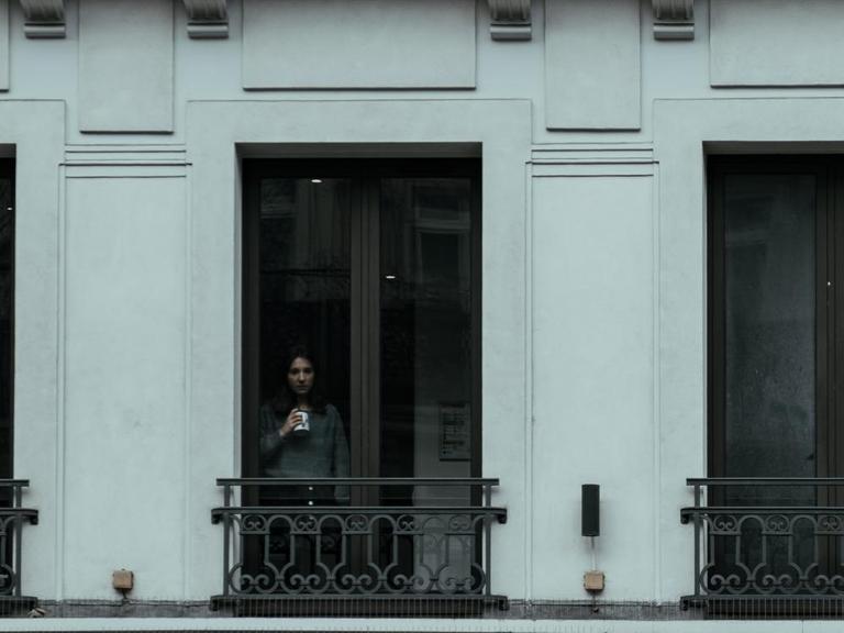 Eine Frau steht mit einer Tasse in der Hand an einem Fenster.