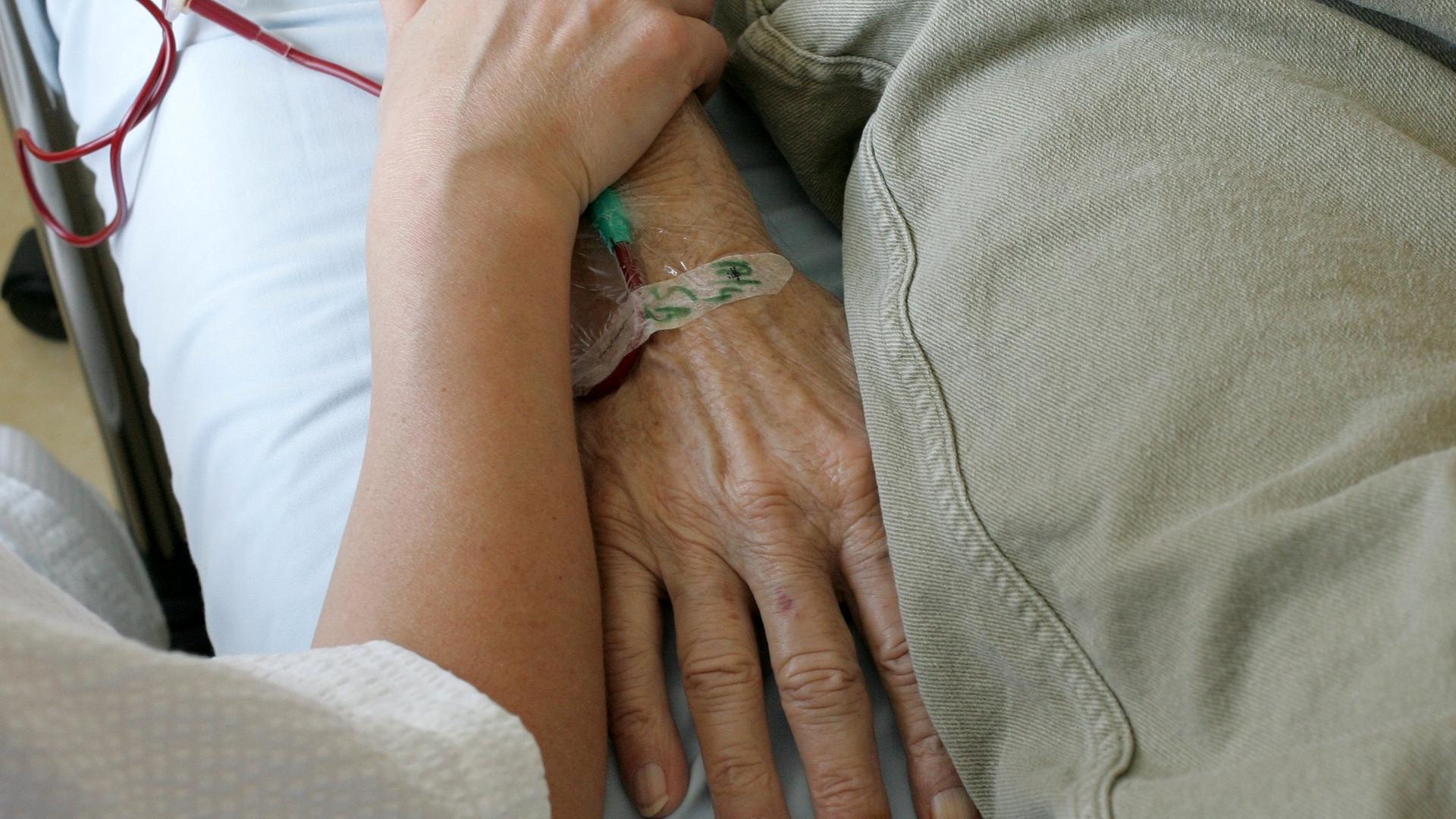 Ein jüngerer Mensch umfasst das Armgelenk einer älteren Person, die im Krankenbett liegt. Ein jüngerer Mensch umfasst das Armgelenk einer älteren Person, die im Krankenbett liegt.