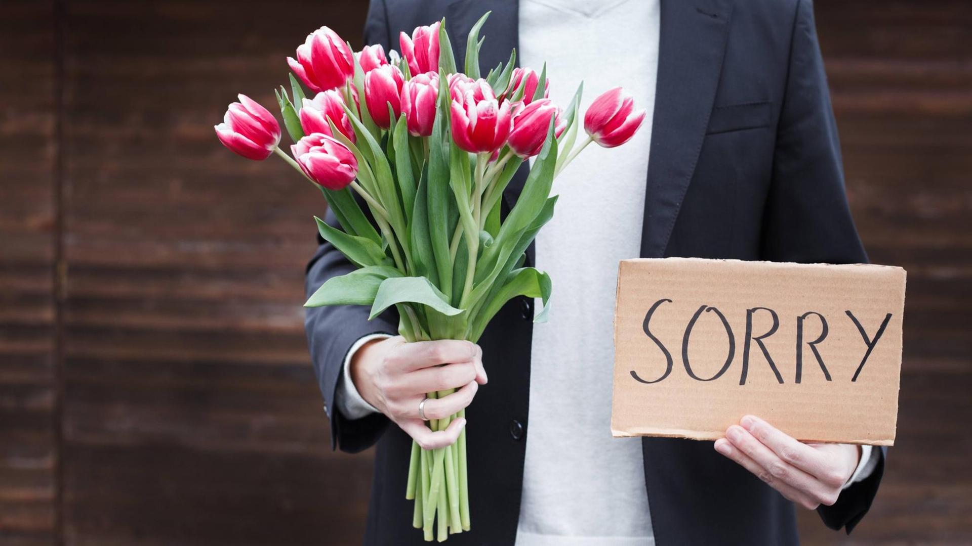 Das Bild zeigt einen Mann, der einen Blumenstrauß und ein Pappschild in den Händen hat. Auf dem Schild steht "sorry".