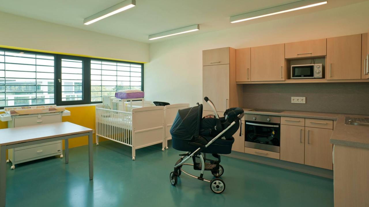 Ein Kinderwagen und Kinderbetten stehen in einem Raum mit vergitterten Fenstern.