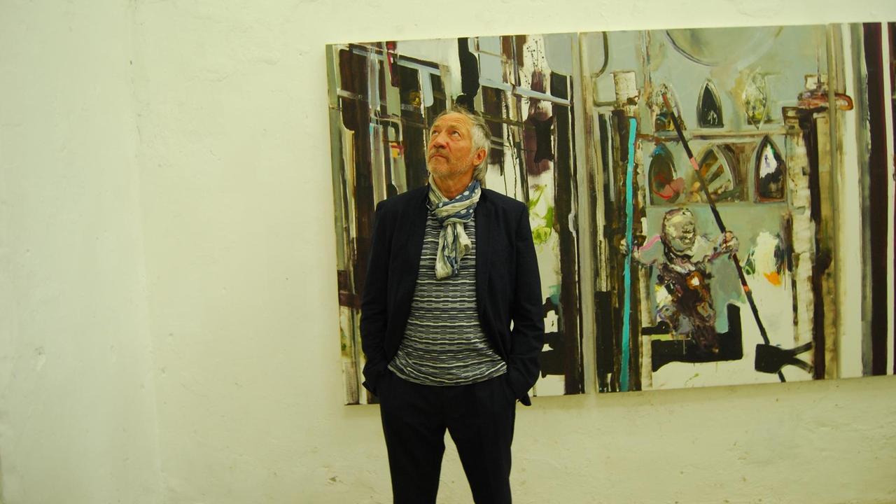 Rudi Kargus, in blauem, legeren Anzug und Turnschuhen vor einem seiner Bilder.