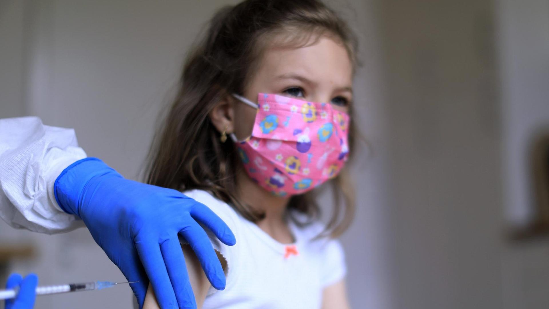Einem Mädchen mit rosa Mund-Nasenschutz wird mit einer Spritze eine Impfung in den Oberarm injiziert. Einem Mädchen mit rosa Mund-Nasenschutz wird mit einer Spritze eine Impfung in den Oberarm injiziert.