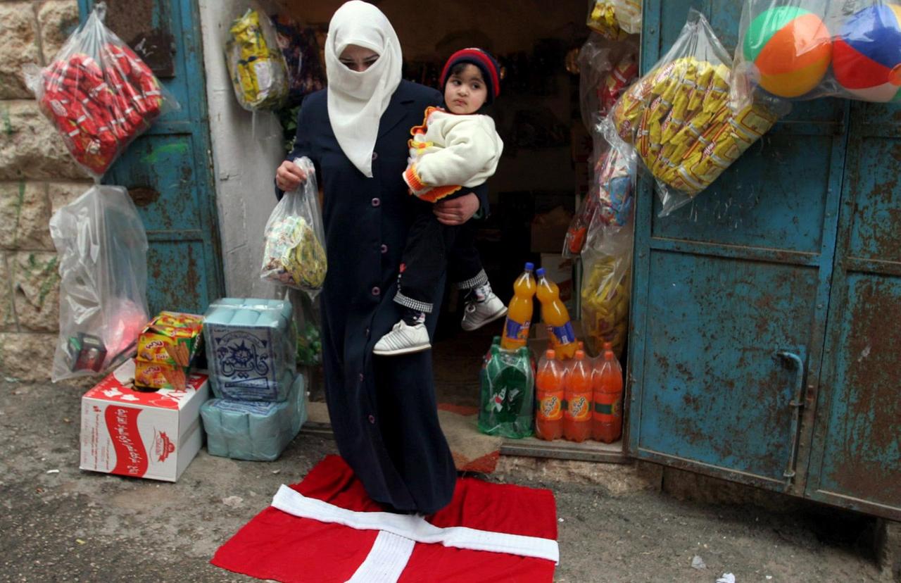Eine Frau in Hebron trägt ihr Kind und ihre Einkäufe über eine dänische Fahne.