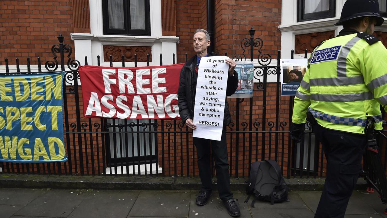 Der Mann steht vor dem Zaun der Botschaft und trägt ein Transparent, auf dem Wikileaks als "Heroes" bezeichnet wird. Ein Polizist läuft an ihm vorbei. Am Zaun hänge Transparente, eines mit der Aufschrift "Free Assange". Der Mann steht vor dem Zaun der Botschaft und trägt ein Transparent, auf dem Wikileaks als "Heroes" bezeichnet wird. Ein Polizist läuft an ihm vorbei. Am Zaun hänge Transparente, eines mit der Aufschrift "Free Assange".