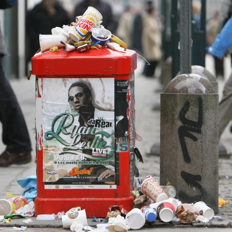 Passanten gehen in der Fußgängerzone in Hamburg an einem überfüllten Abfalleimer vorbei.
