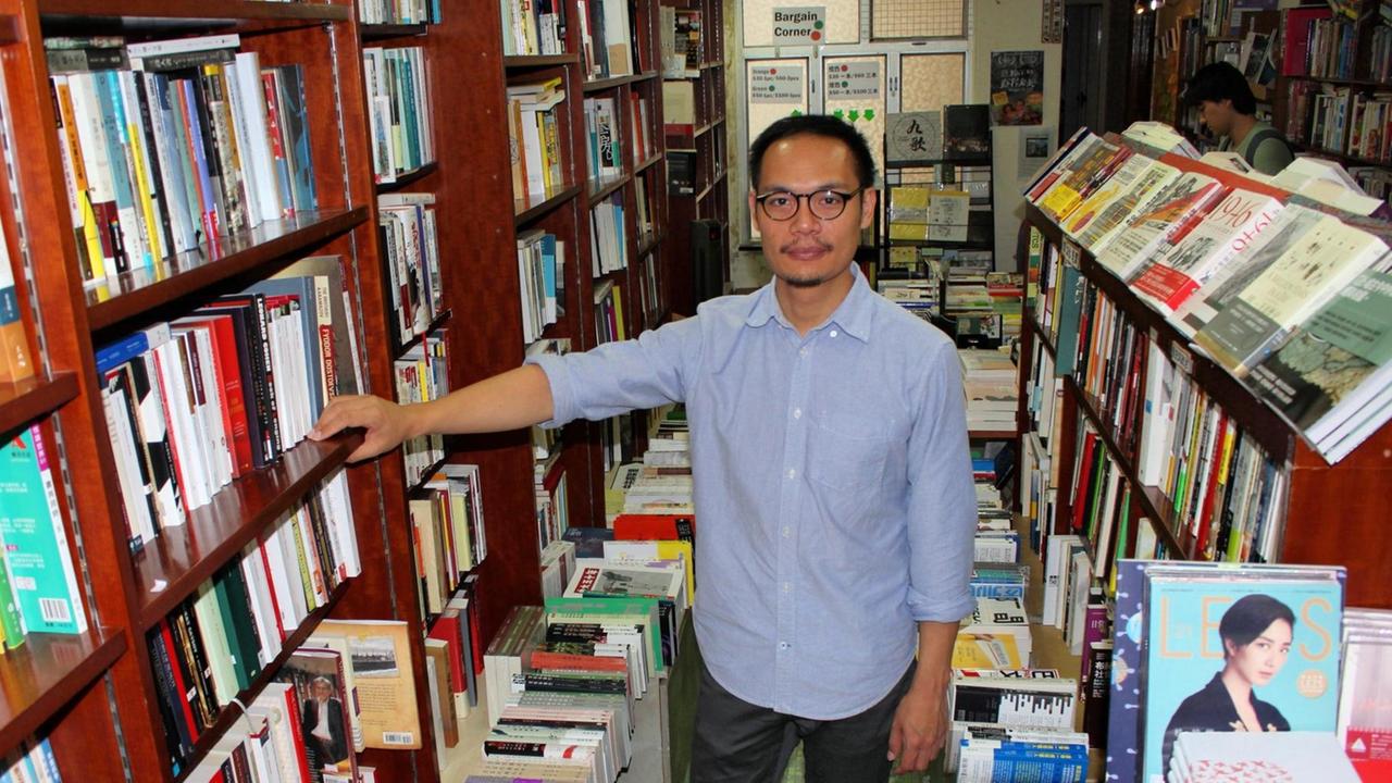 Daniel Lee, Buchhändler in Hongkong, steht im hellblauen Hemd, mit Brille, in seinem Laden inmitten voller Bücherregale und blickt in die Kamera. Daniel Lee, Buchhändler in Hongkong, steht im hellblauen Hemd, mit Brille, in seinem Laden inmitten voller Bücherregale und blickt in die Kamera.