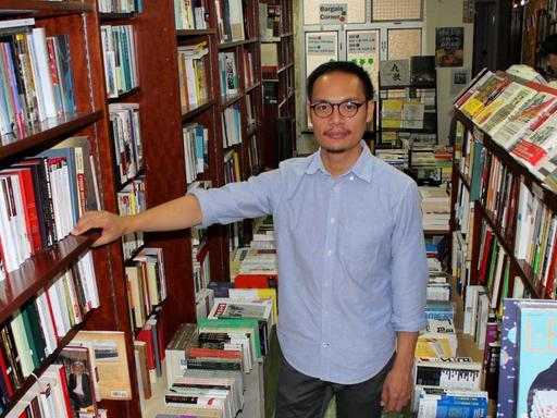 Daniel Lee, Buchhändler in Hongkong, steht im hellblauen Hemd, mit Brille, in seinem Laden inmitten voller Bücherregale und blickt in die Kamera.