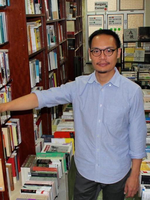 Daniel Lee, Buchhändler in Hongkong, steht im hellblauen Hemd, mit Brille, in seinem Laden inmitten voller Bücherregale und blickt in die Kamera.