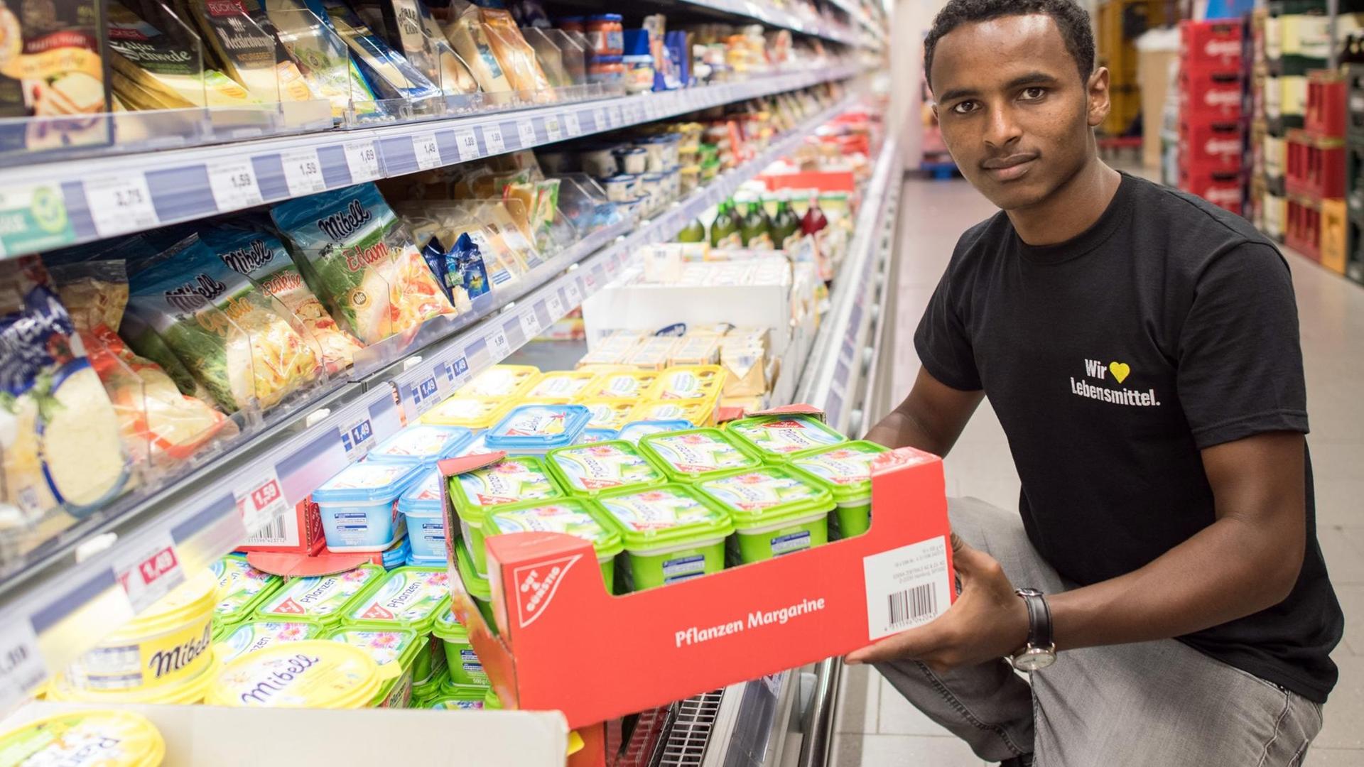 Ein junger Geflüchteter räumt ein Kühlregal im Supermarkt ein und blickt dabei in die Kamera. Ein junger Geflüchteter räumt ein Kühlregal im Supermarkt ein und blickt dabei in die Kamera.