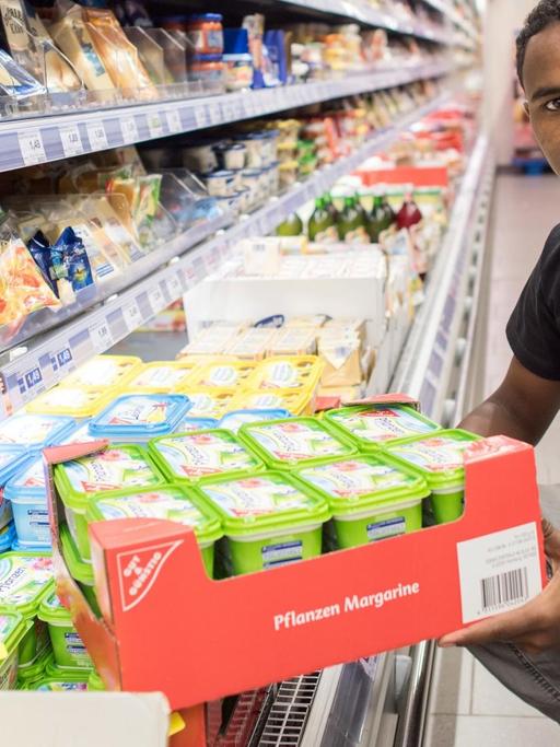 Ein junger Geflüchteter räumt ein Kühlregal im Supermarkt ein und blickt dabei in die Kamera.