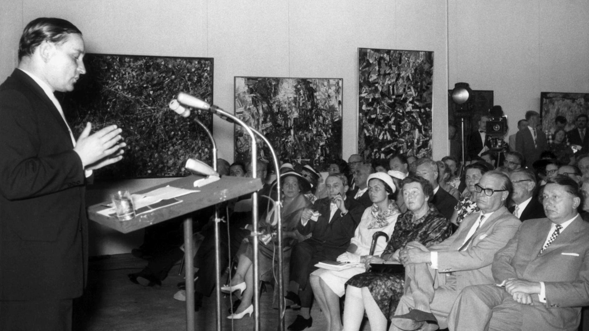 Der Kunsthistoriker Werner Haftmann steht während der Eröffnung der documenta 2 im Jahr 1959 am Rednerpult, im Publikum unter anderem sein Kollege Arnold Bode.