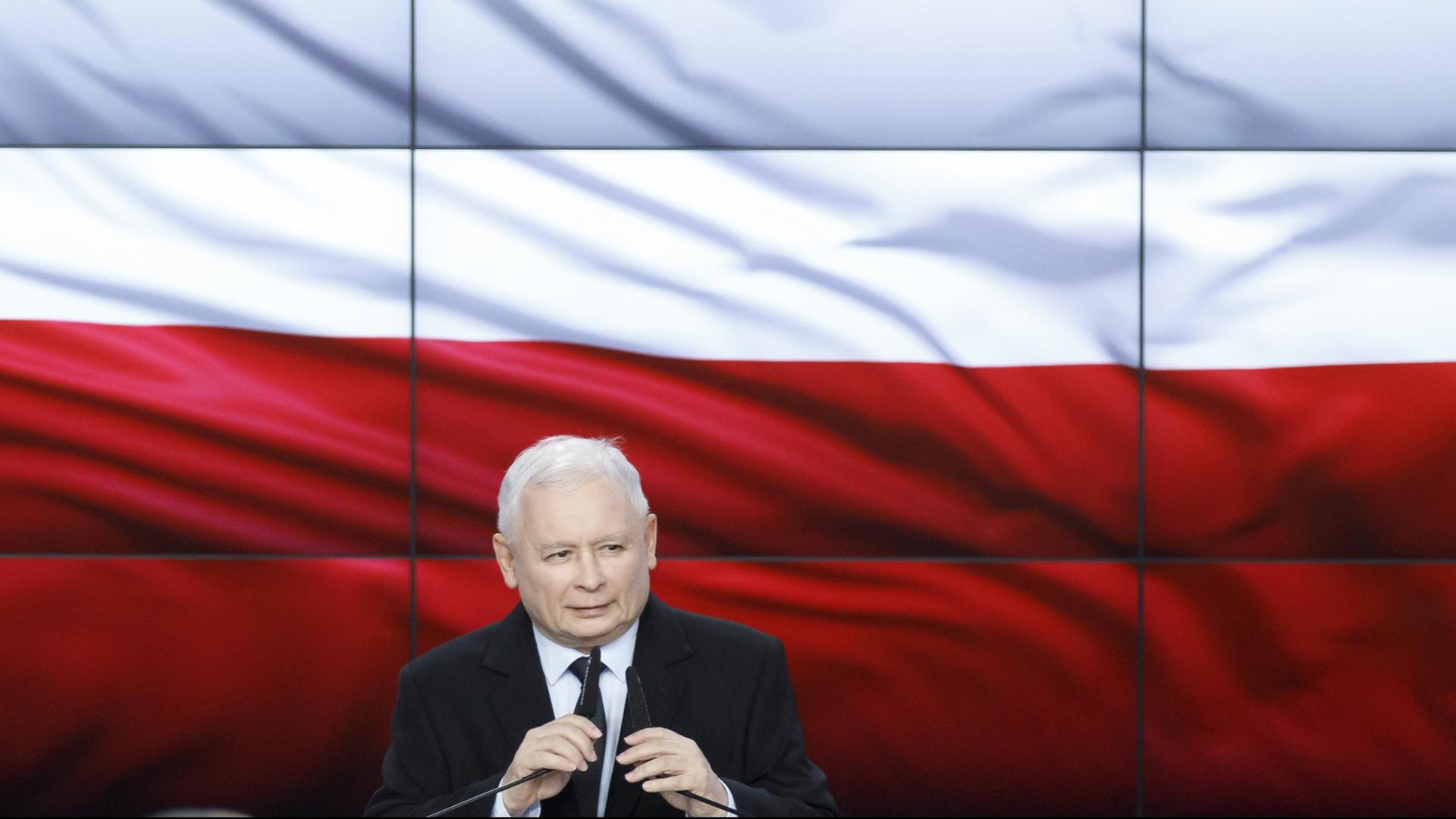 Jaroslaw Kaczynski hinter zwei Mikrofonen, im Hintergrund die polnischen Landesfarben