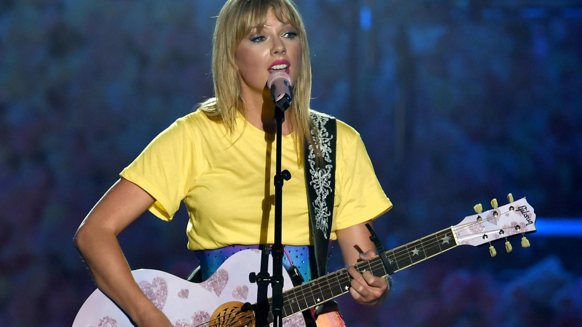 Das Bild zeigt Taylor Swift während eines Konzerts wie sie singt und dabei auf einer rosa Gitarre spielt. Das Bild zeigt Taylor Swift während eines Konzerts wie sie singt und dabei auf einer rosa Gitarre spielt.