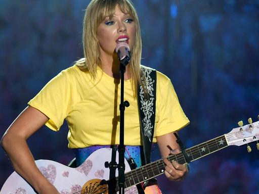 Das Bild zeigt Taylor Swift während eines Konzerts wie sie singt und dabei auf einer rosa Gitarre spielt.