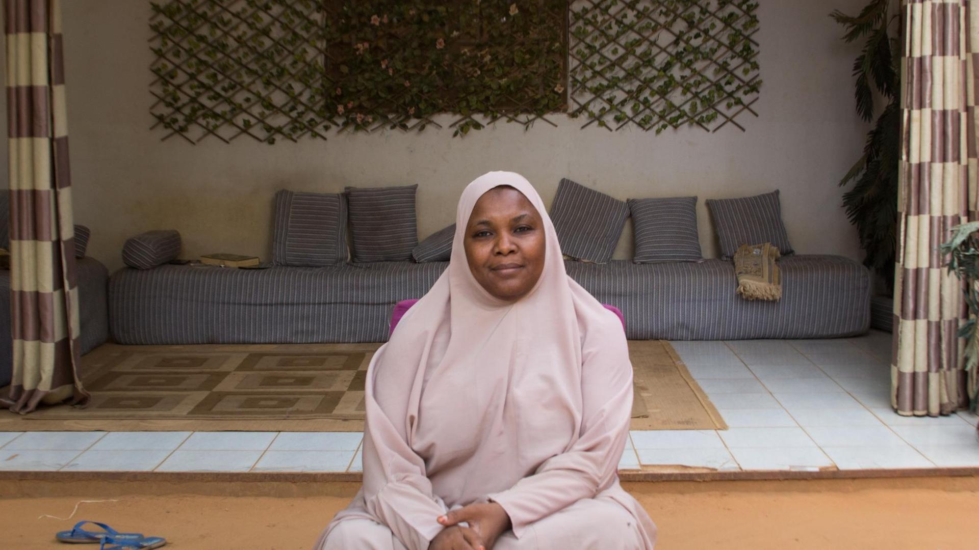 Die Predigerin Malama Sakina, in mauvefarbenes Tuch und Kopftuch gehüllt, sitzt in ihrem Haus vor einer mit Vorhängen gerahmten Nische, in der unter dem Fenster ein Sofa mit vielen kleinen Kissen steht.