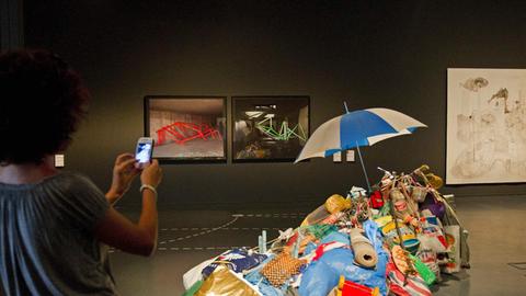 Eine Besucherin macht ein Foto von einem Ausstellungsstück von Andreas Slominski. Zu sehen sind gefüllte Müllsäcke, Tüten, Beutel, Alltagsgegenstände wie einen Teppich und Spielzeug, die auf einem Haufen liegen. Ganz oben steckt ein Regeschirm. Das Ganze ist der Teil der Ausstellung, die 2013 in Barcelona gezeigt wurde.