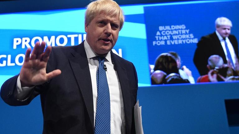 Johnson läuft vor der blau erstrahlenden Bühne entlang und winkt in die Kamera.