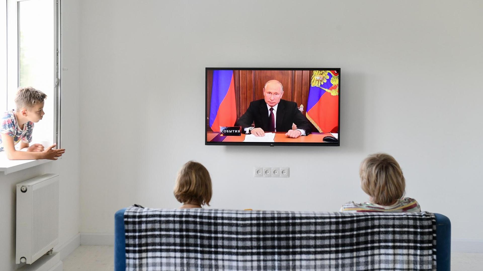 Der russische Präsident Wladimir Putin hält eine Ansprache im Fernsehen. Zwei russische Bürgerinnen sitzen auf einem Sofa und hören zu.