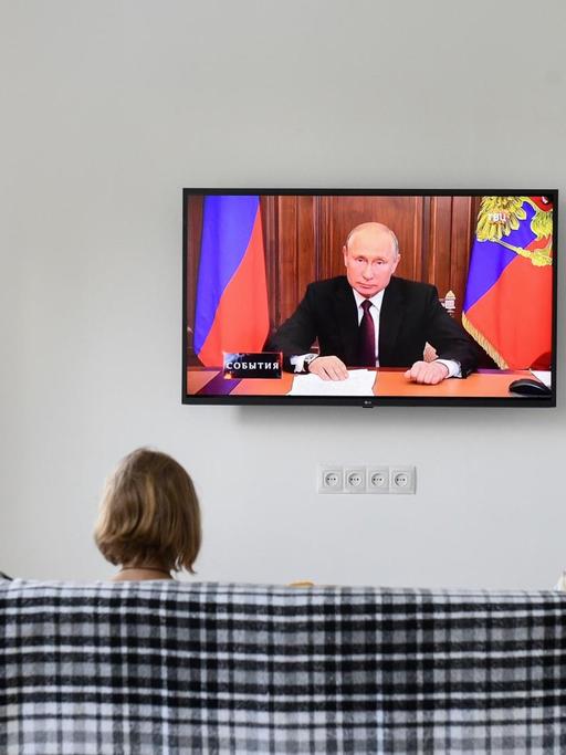 Der russische Präsident Wladimir Putin hält eine Ansprache im Fernsehen. Zwei russische Bürgerinnen sitzen auf einem Sofa und hören zu.