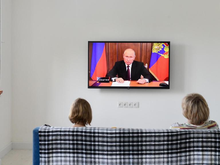 Der russische Präsident Wladimir Putin hält eine Ansprache im Fernsehen. Zwei russische Bürgerinnen sitzen auf einem Sofa und hören zu.