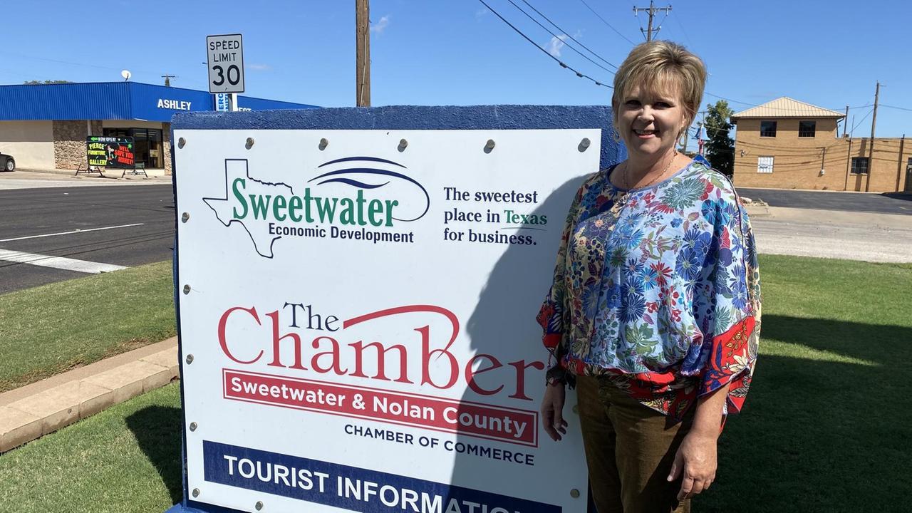 Eine blonde Frau in bunter Bluse steht vor einem Schild der Tourismusinformation von Sweetwater.