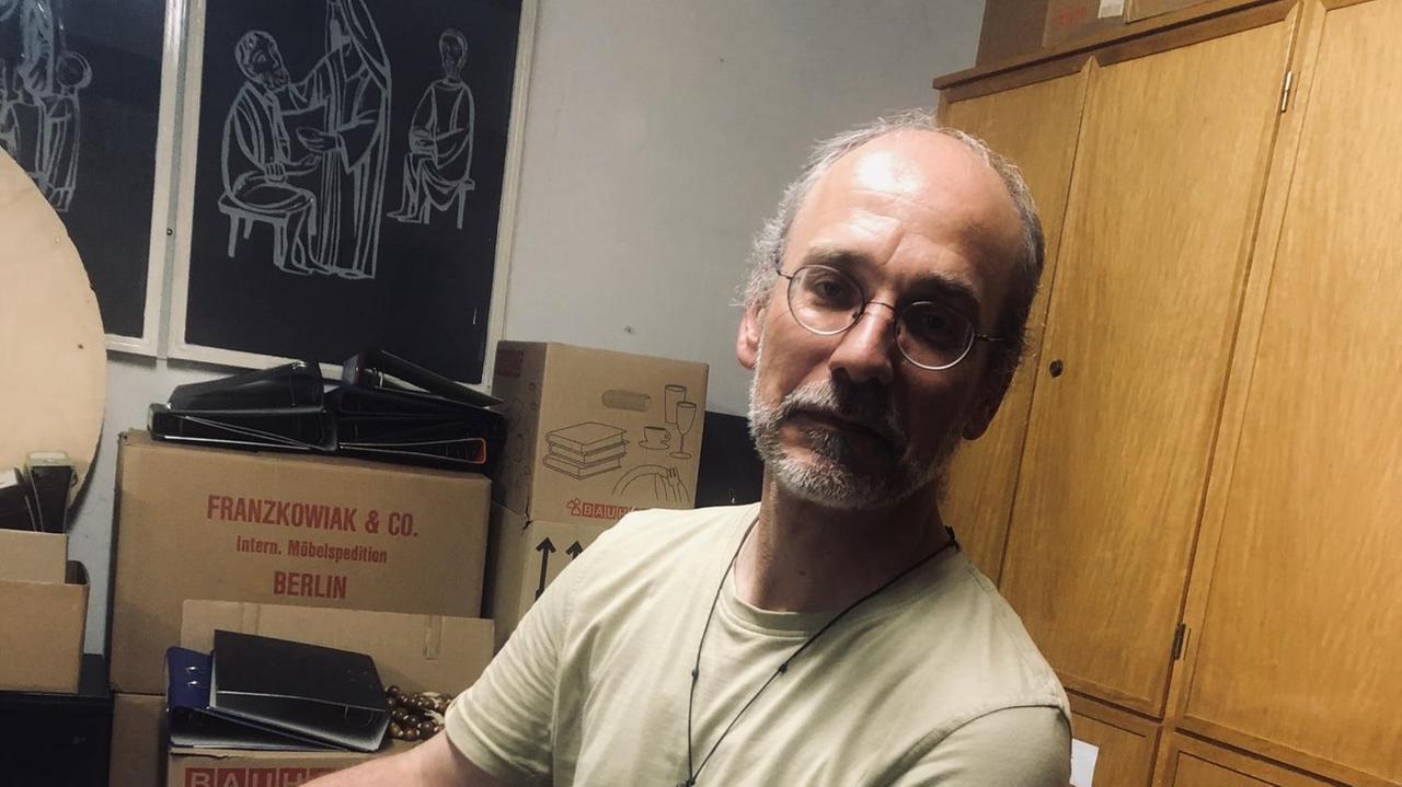 Pater Damian Bieger im Archiv der Gemeinde St. Ludwig, umgeben von Umzugskartons und Aktenordnern. Auf einem Bild im Hintergrund: das religiöse Motiv einer Frau, die einem Mann zu essen gibt. Pater Damian Bieger im Archiv der Gemeinde St. Ludwig, umgeben von Umzugskartons und Aktenordnern. Auf einem Bild im Hintergrund: das religiöse Motiv einer Frau, die einem Mann zu essen gibt.