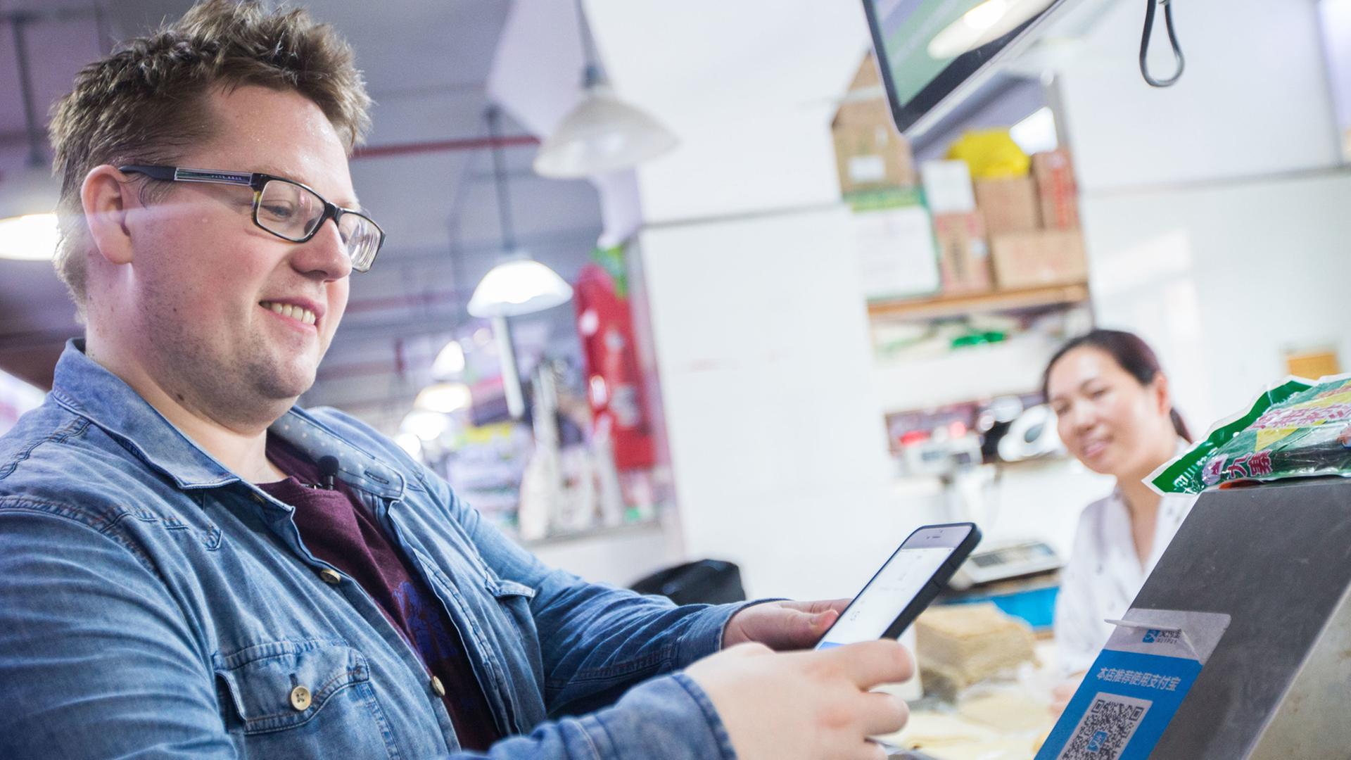 Das Bild zeigt einen jungen Mann mit Bürstenschnitt, Jeansjacke und Brille, der mit seinem Smartphone in einem Supermarkt in Shanghai einen QR Code scannt. Das Bild zeigt einen jungen Mann mit Bürstenschnitt, Jeansjacke und Brille, der mit seinem Smartphone in einem Supermarkt in Shanghai einen QR Code scannt.