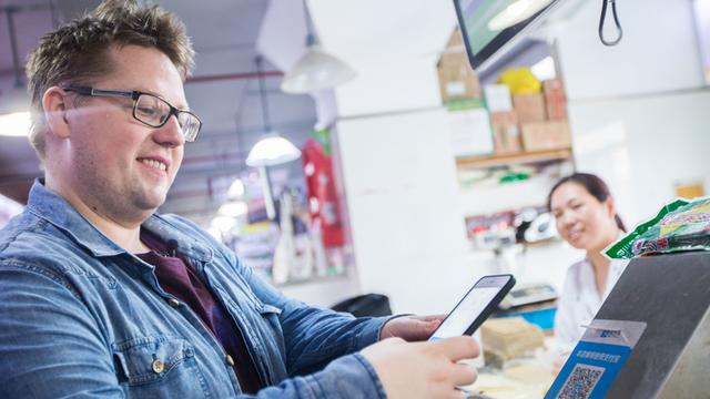 Das Bild zeigt einen jungen Mann mit Bürstenschnitt, Jeansjacke und Brille, der mit seinem Smartphone in einem Supermarkt in Shanghai einen QR Code scannt.