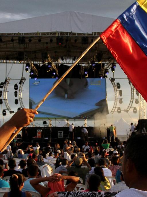 Ein Mann schwenkt eine kolumbianische Fahne. Im Hintergrund ist eine Bühne zu sehen.