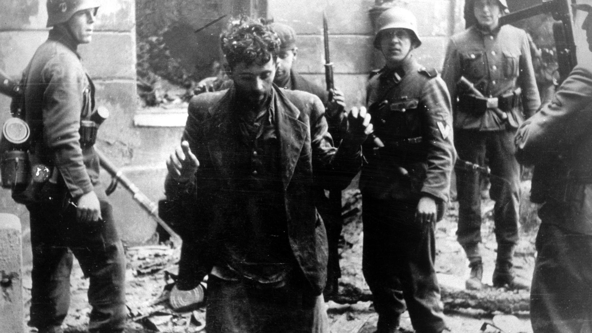 Zwei Juden, die sich in einem Haus versteckt hatten, werden von SS-Soldaten gefangen genommen. Die Aufnahme entstand während des Warschauer Ghetto-Aufstands, der vom 19. April 1943 bis zu seiner blutigen Niederschlagung am 16. Mai 1943 dauerte. Die Nationalsozialisten hatten ein Jahr nach der Besetzung Polens im November 1940 in Warschau ein Ghetto errichtet und dorthin annähernd eine halbe Million Juden verschleppt. Zwischen Juli und September 1942 wurden 300 000 Opfer in den Todeslagern, die meisten in Treblinka, ermordet. Als am 19. April 1943 die SS mit der Verschleppung der restlichen 60 000 Ghetto-Einwohner begann, leisteten mehrere hundert militärisch organisierte Juden bewaffneten Widerstand. Bei den Kämpfen wurden etwa 14 000 jüdische Aufständische getötet. Zwei Juden, die sich in einem Haus versteckt hatten, werden von SS-Soldaten gefangen genommen. Die Aufnahme entstand während des Warschauer Ghetto-Aufstands, der vom 19. April 1943 bis zu seiner blutigen Niederschlagung am 16. Mai 1943 dauerte. Die Nationalsozialisten hatten ein Jahr nach der Besetzung Polens im November 1940 in Warschau ein Ghetto errichtet und dorthin annähernd eine halbe Million Juden verschleppt. Zwischen Juli und September 1942 wurden 300 000 Opfer in den Todeslagern, die meisten in Treblinka, ermordet. Als am 19. April 1943 die SS mit der Verschleppung der restlichen 60 000 Ghetto-Einwohner begann, leisteten mehrere hundert militärisch organisierte Juden bewaffneten Widerstand. Bei den Kämpfen wurden etwa 14 000 jüdische Aufständische getötet.