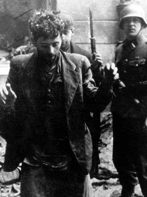 Zwei Juden, die sich in einem Haus versteckt hatten, werden von SS-Soldaten gefangen genommen. Die Aufnahme entstand während des Warschauer Ghetto-Aufstands, der vom 19. April 1943 bis zu seiner blutigen Niederschlagung am 16. Mai 1943 dauerte. Die Nationalsozialisten hatten ein Jahr nach der Besetzung Polens im November 1940 in Warschau ein Ghetto errichtet und dorthin annähernd eine halbe Million Juden verschleppt. Zwischen Juli und September 1942 wurden 300 000 Opfer in den Todeslagern, die meisten in Treblinka, ermordet. Als am 19. April 1943 die SS mit der Verschleppung der restlichen 60 000 Ghetto-Einwohner begann, leisteten mehrere hundert militärisch organisierte Juden bewaffneten Widerstand. Bei den Kämpfen wurden etwa 14 000 jüdische Aufständische getötet.