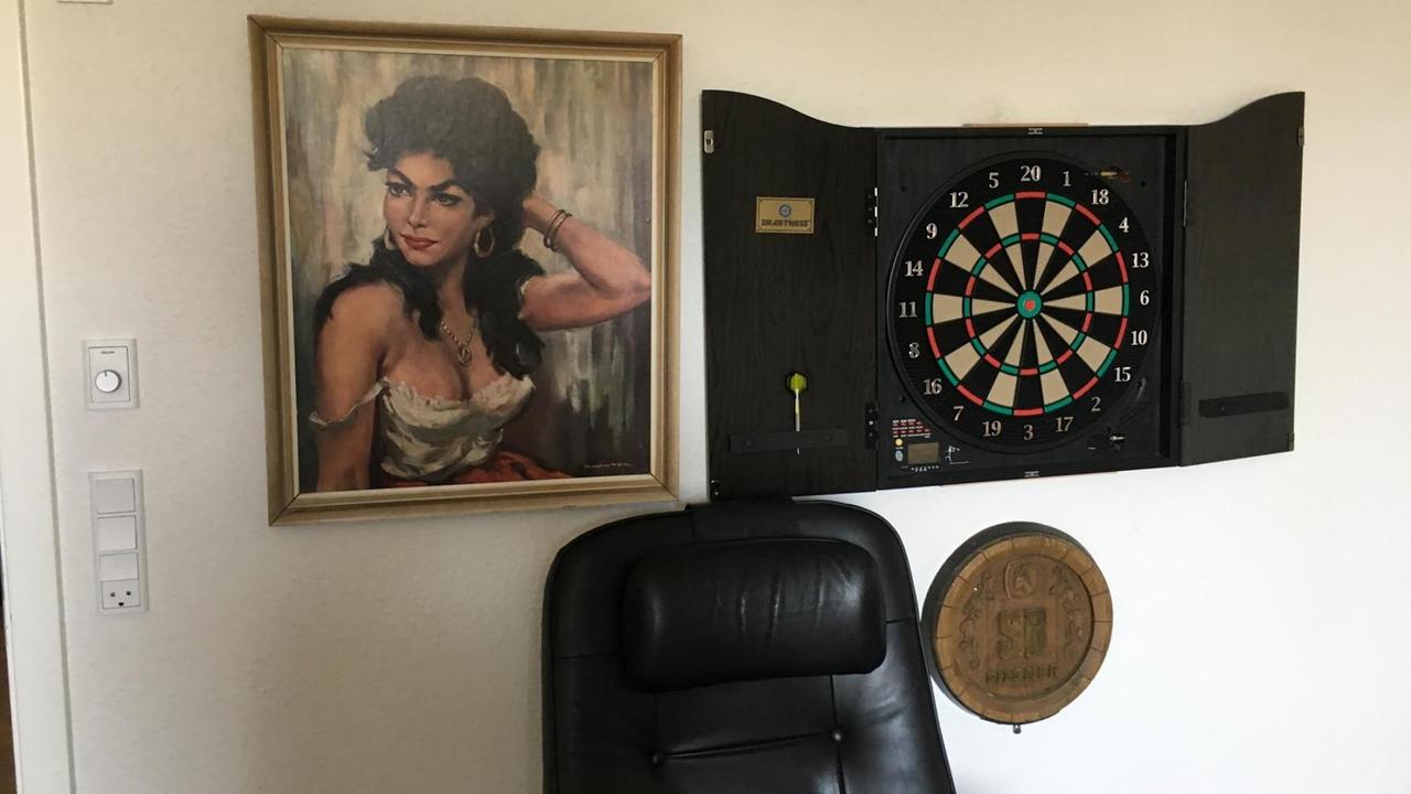 Ein Zimmer mit einem schwarzen Sessel, dem Gemälde einer Zigeunerin und einer Dartscheibe. Ein Zimmer mit einem schwarzen Sessel, dem Gemälde einer Zigeunerin und einer Dartscheibe.