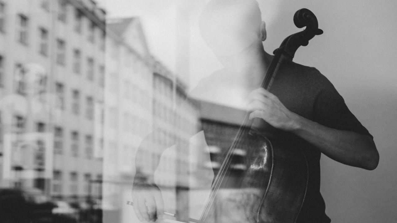 Ein Cellist spielt hinter einer Fensterscheibe, in der sich ein historischer Straßenzug spiegelt.