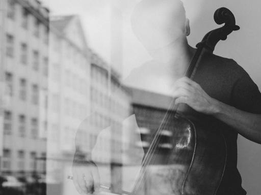 Ein Cellist spielt hinter einer Fensterscheibe, in der sich ein historischer Straßenzug spiegelt.