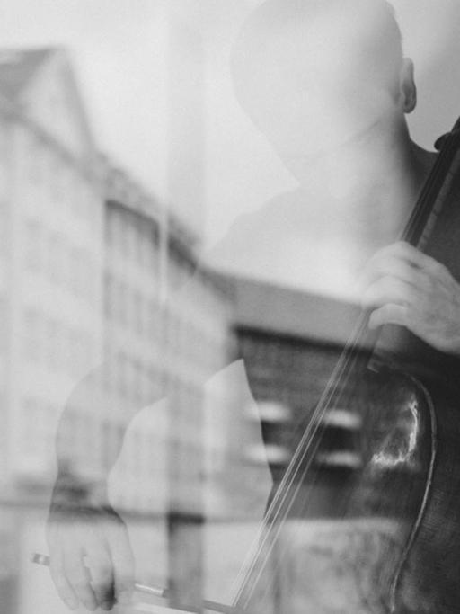 Ein Cellist spielt hinter einer Fensterscheibe, in der sich ein historischer Straßenzug spiegelt.