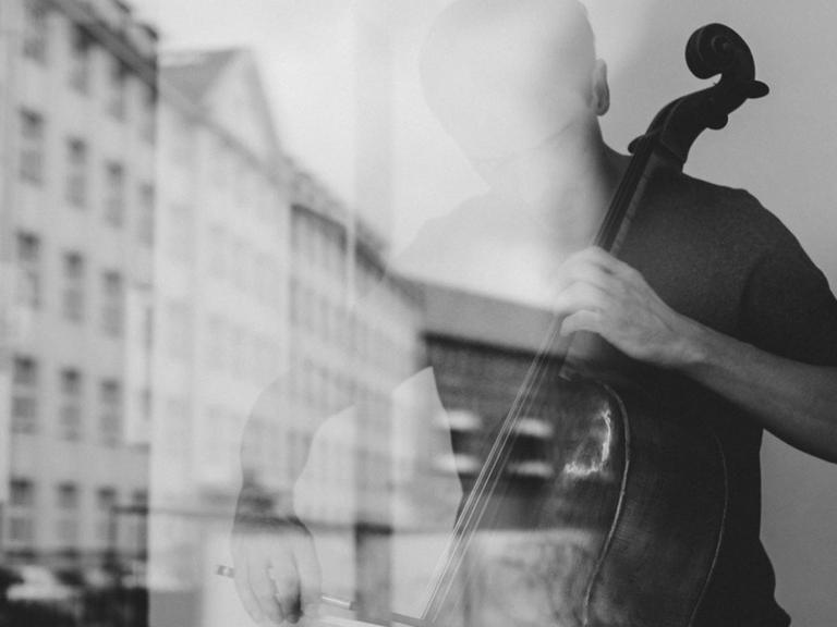 Ein Cellist spielt hinter einer Fensterscheibe, in der sich ein historischer Straßenzug spiegelt.
