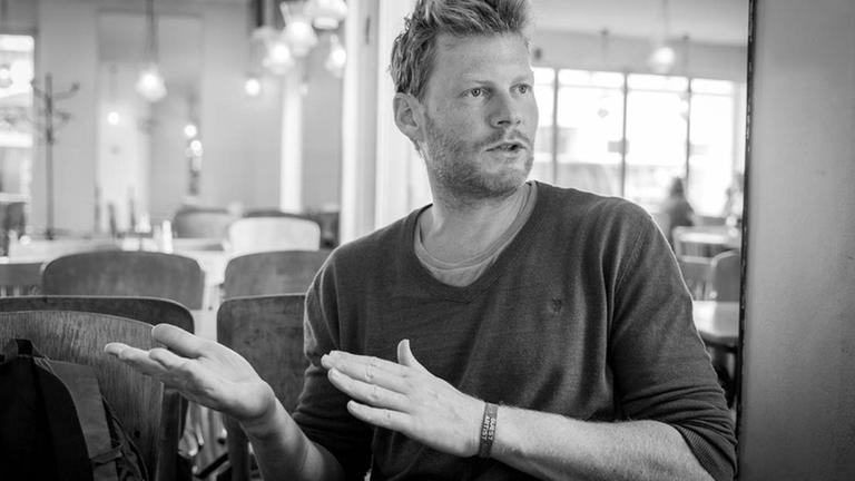 Schwarzweiß-Aufnahme von Christian Felber. Er sitzt gestikulieren und im Gespräch in einem Cafe. Schwarzweiß-Aufnahme von Christian Felber. Er sitzt gestikulieren und im Gespräch in einem Cafe.