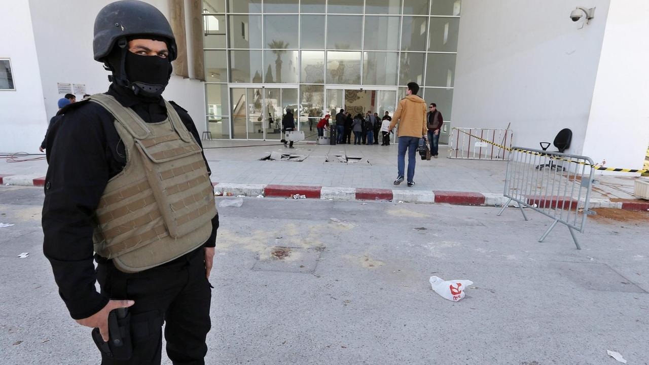 Ein schwarz gekleideter Polizist mit Helm, Sturmhaube und kugelsicherer Weste steht vor dem abgesperrten Eingang des Bardo-Museums in Tunis. Hinter ihm sieht man Ermittler; rechts vom Eingang sind Blutspuren zu sehen. 