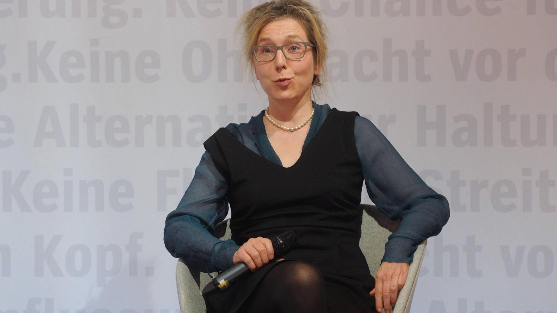 Elke Schmitter sitzt auf einem Sessel auf einem Podium und hält ein Mikrofon in der Hand
